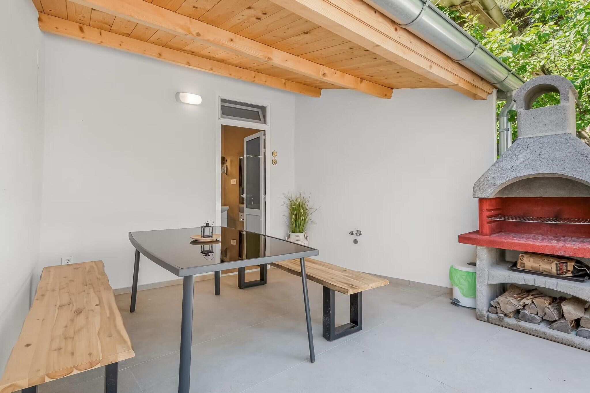 Een moderne eethoek buiten met een glazen tafel, houten banken en een steenoven voor pizza.
