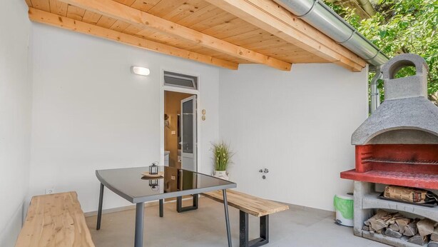 Un área de comedor moderna al aire libre con mesa de vidrio, bancos de madera y un horno de pizza de piedra.