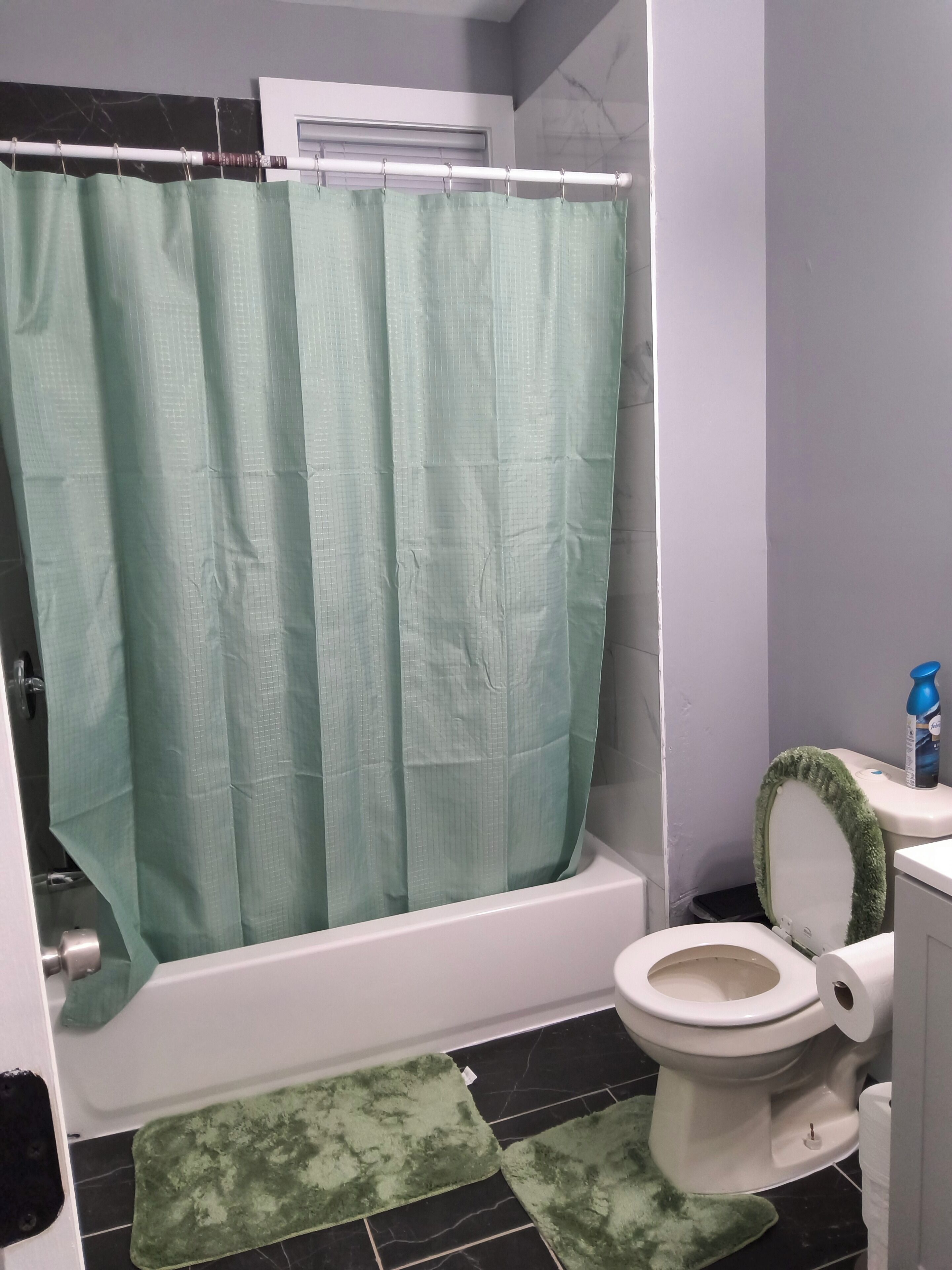 Un baño con cortina de ducha verde, un inodoro con tapete verde y una botella azul sobre el lavamanos.