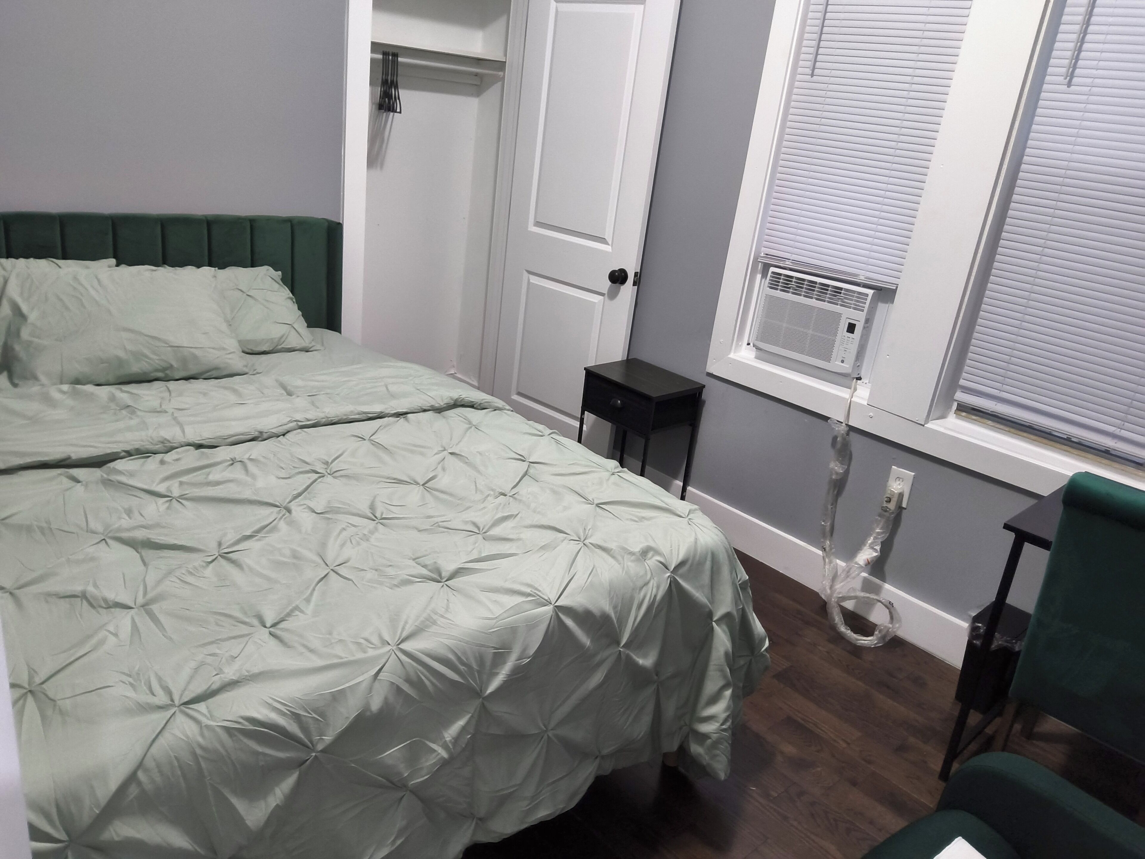 A bedroom with a bed, a nightstand, a chair, a window with blinds, and a door.