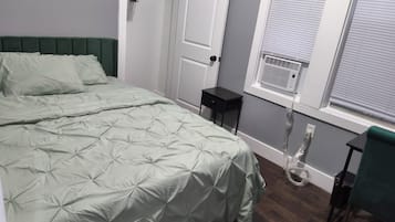A bedroom with a bed, a nightstand, a chair, a window with blinds, and a door.