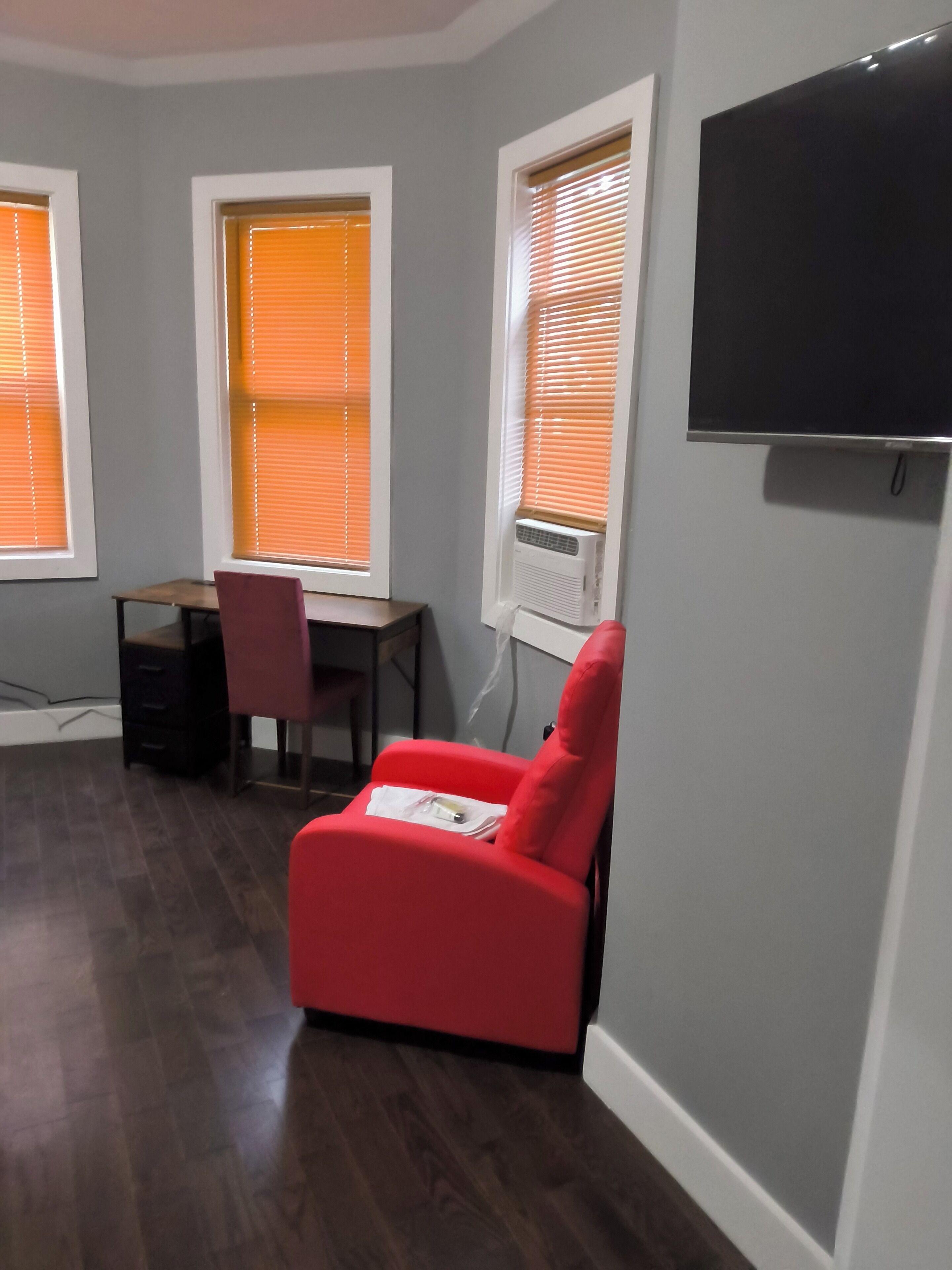 A room with a red chair, a desk, and a television mounted on the wall.