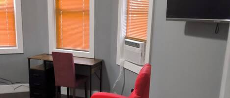 A room with a red chair, a desk, and a television mounted on the wall.