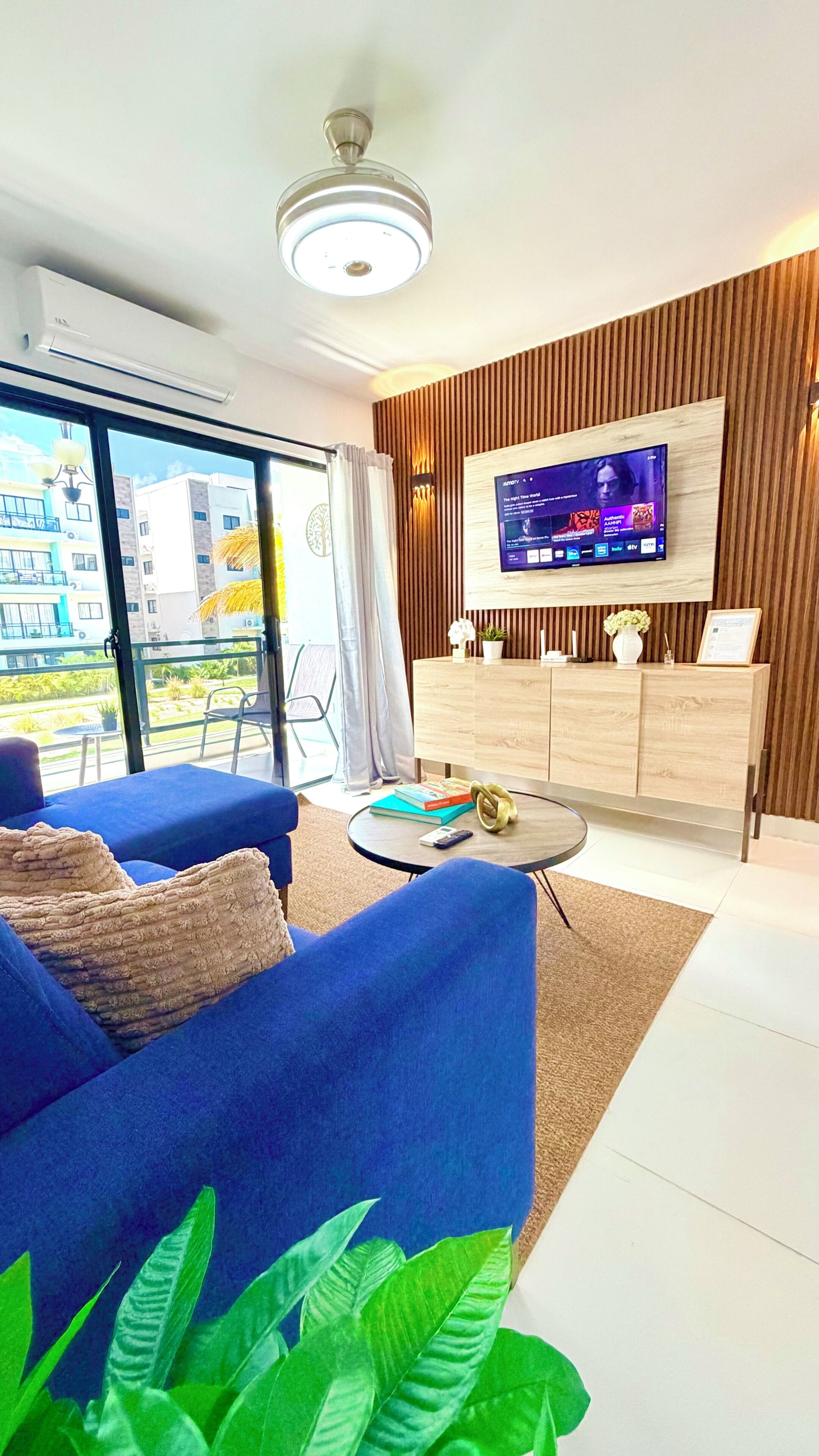 A modern living room with a blue sofa, a TV mounted on a wooden wall, a round coffee table, and a view of buildings outside.