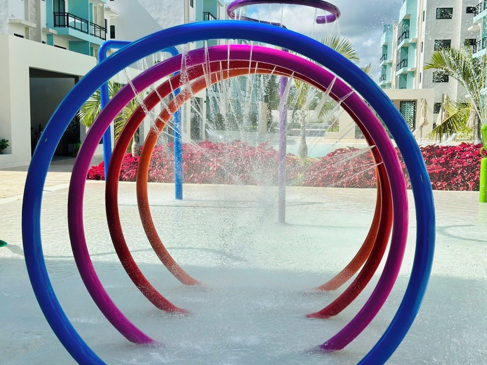 Ein Wasserspiel mit mehreren Ringen, umgeben von einem modernen Gebäude mit Balkonen und roten Blumen.