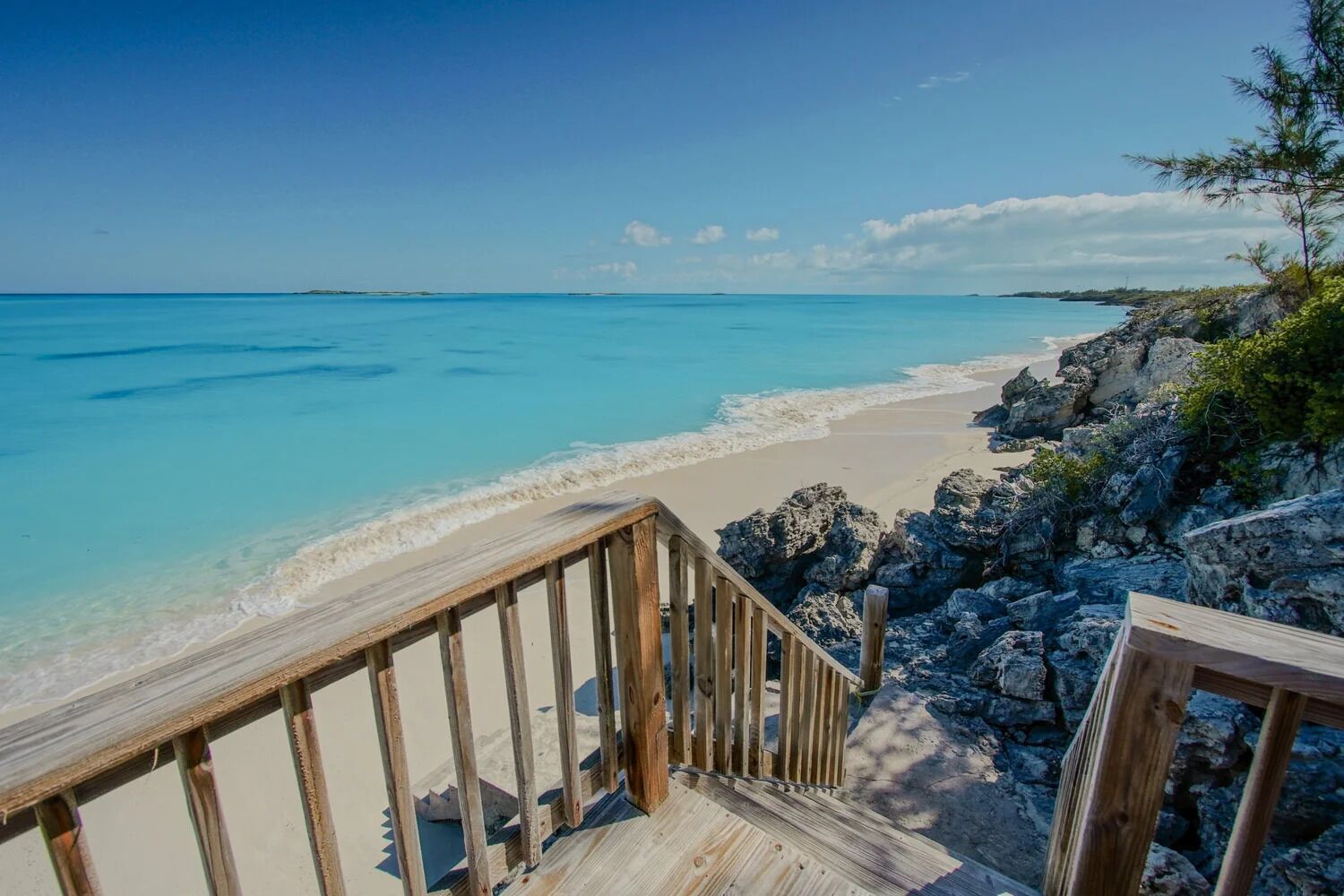A wooden staircase leading to a beach with clear turquoise water and rocky terrain.