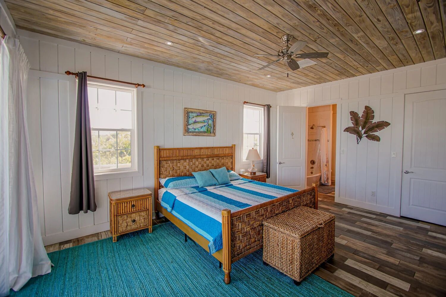 A bedroom with a wooden ceiling, a bed with a blue and white striped bedspread, a wicker bench, and a painting on the wall.
