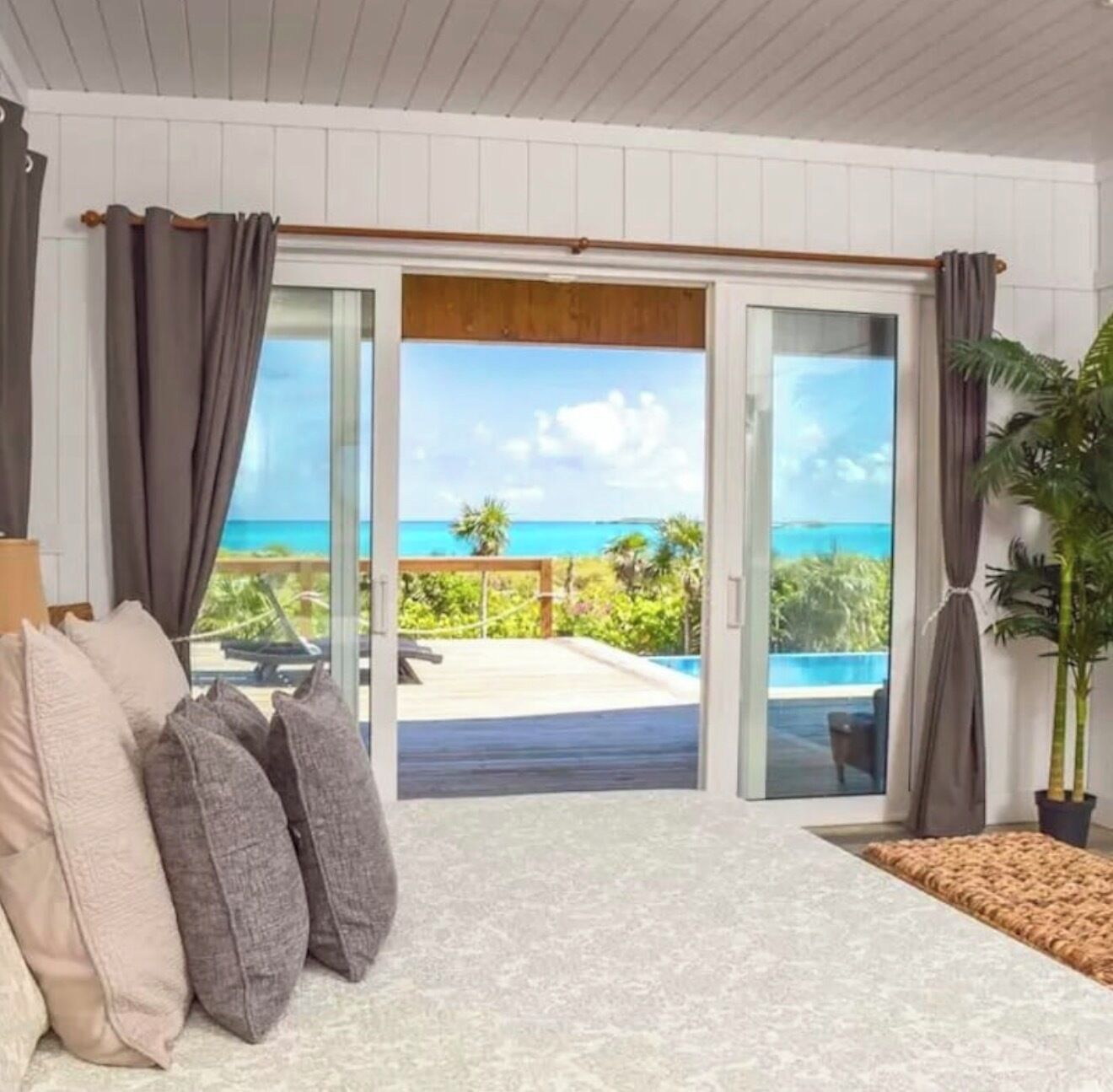A room with a view of the ocean, a sliding glass door, a bed with pillows, and a potted plant.