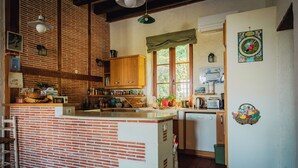 Eine Küche mit Kochinsel aus Backstein, Holzschränken, einem Fenster mit kariertem Vorhang und einem Kühlschrank mit Obstschale.