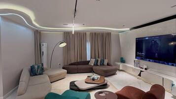 Una sala de estar moderna con un televisor de pantalla plana grande, un sofá marrón, un pouf color turquesa y una mesa de centro.