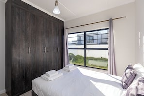 A bedroom with a large wardrobe, a bed with white linens, and a view through a window. - Modern 3-Bed Garden Pool Gym by CTHA (Cape Town)