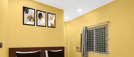 A hotel room with a bed, a window with a metal grill, and three framed artworks on the wall.