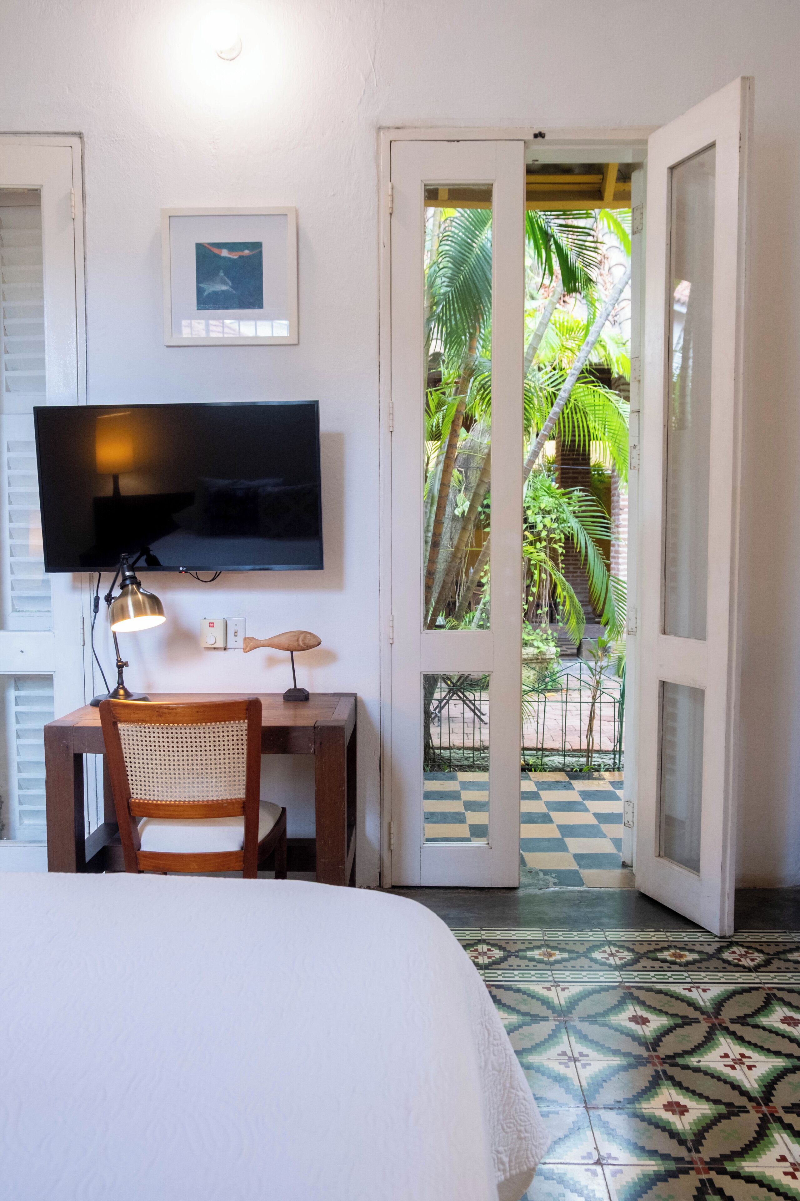 A bedroom with a bed, a desk with a chair, a television, and a door leading to a balcony with a view of greenery.