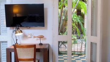 A bedroom with a bed, a desk with a chair, a television, and a door leading to a balcony with a view of greenery.