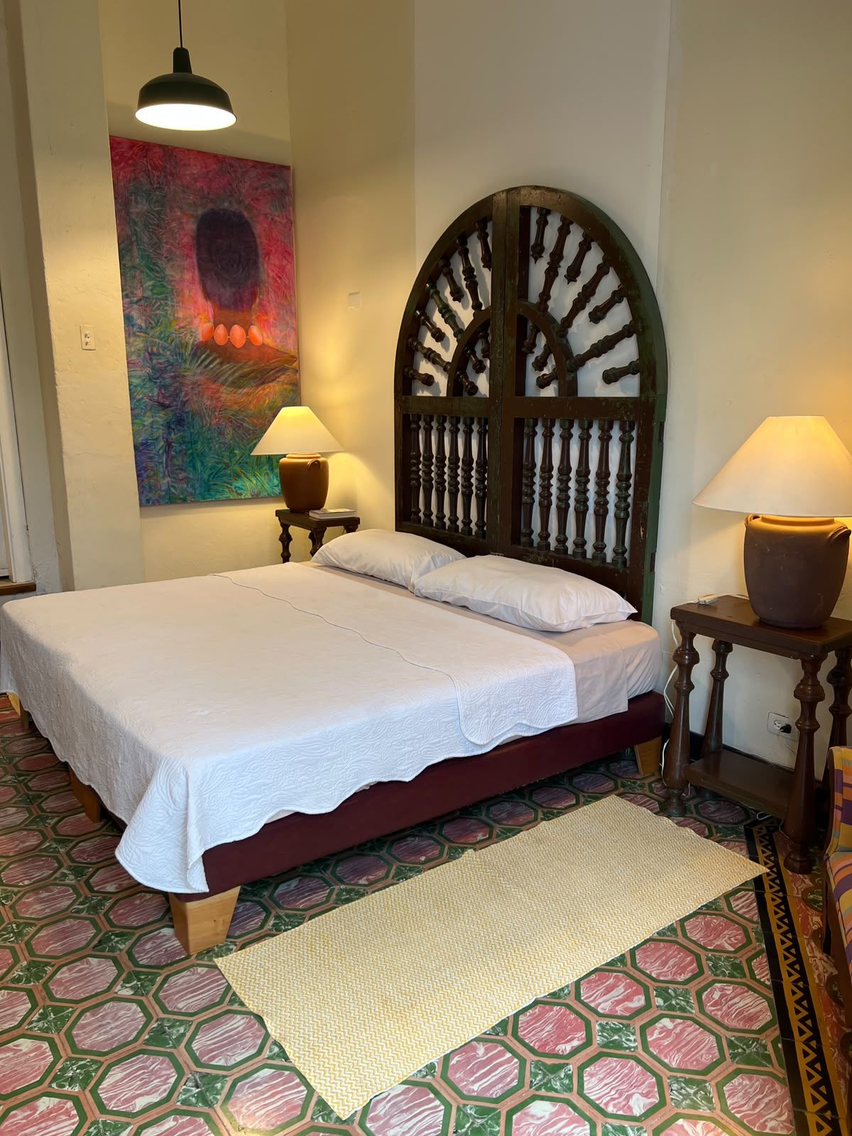 A bedroom with a bed, two bedside lamps, a decorative headboard, a colorful abstract painting, and patterned flooring.