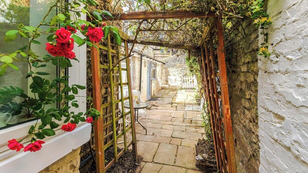 Un jardín con un enrejado de madera, rosas rojas y un sendero de piedra que lleva a un edificio blanco.