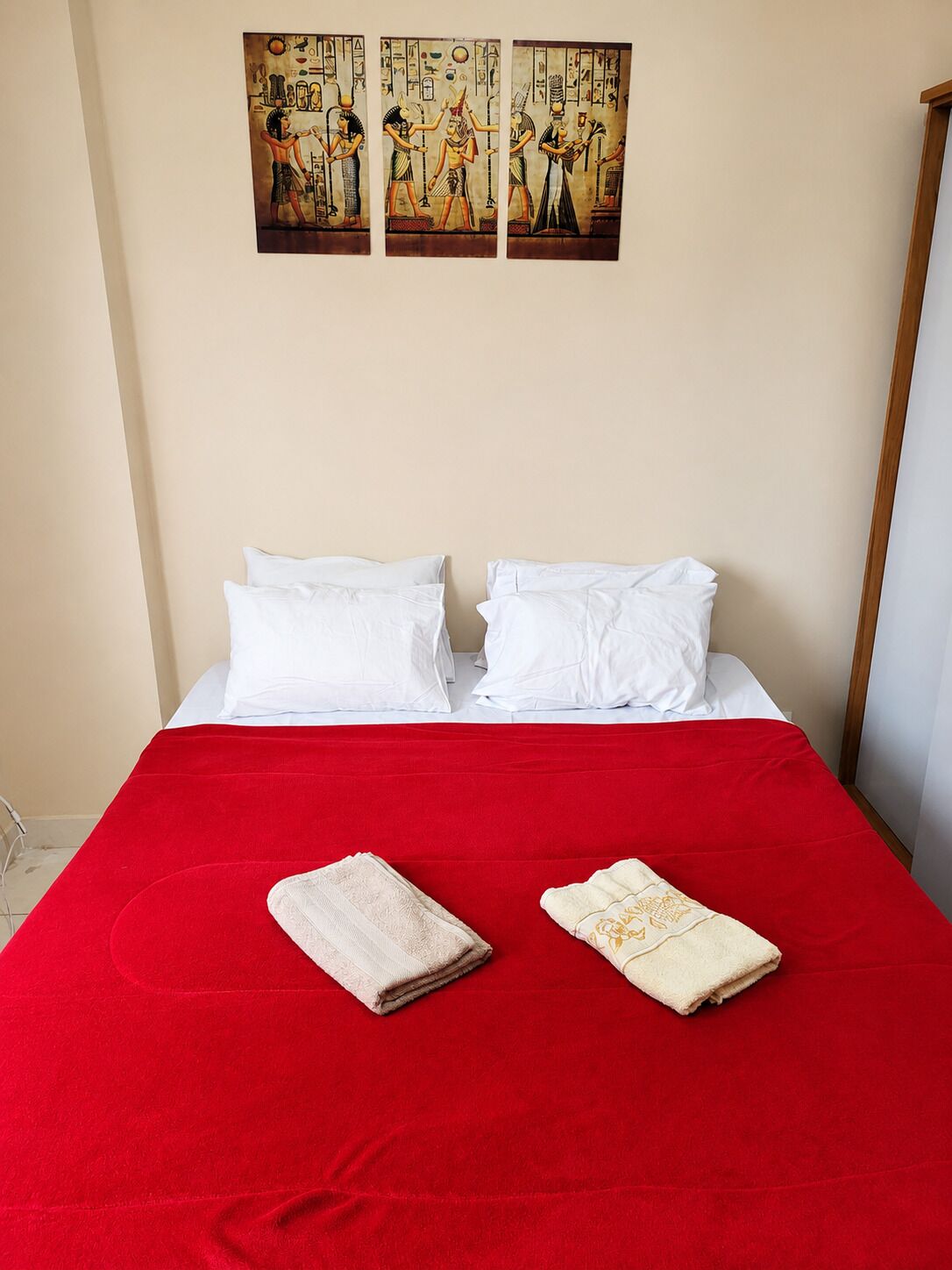 Uma cama com almofadas brancas e um cobreleito vermelho, duas toalhas dobradas e três quadros na parede acima da cama.