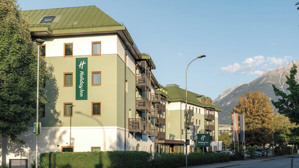 Um hotel Holiday Inn com várias andares, varandas, telhado verde e montanhas ao fundo.