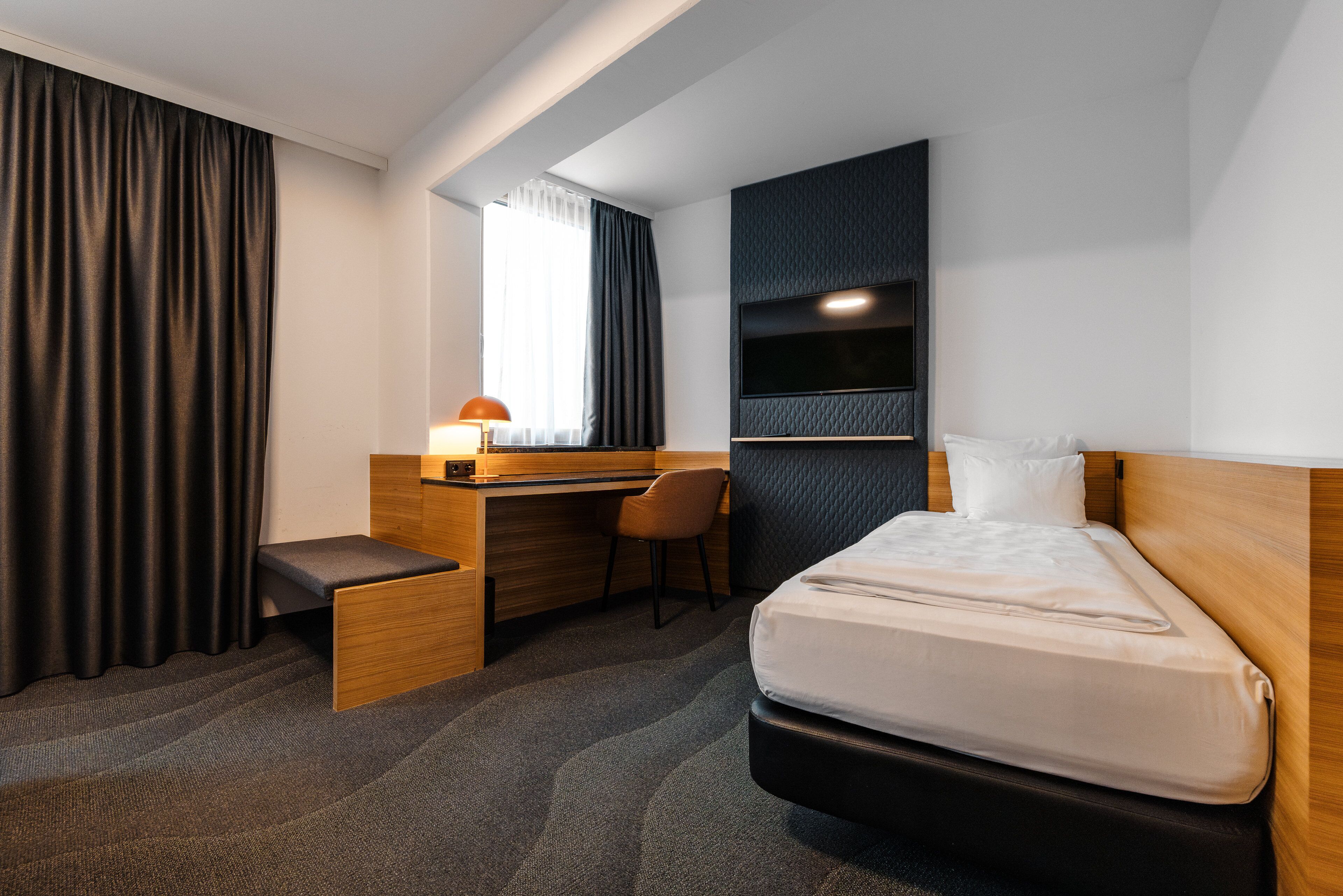 Una habitación de hotel con cama, escritorio, silla, televisor y ventana con cortinas.