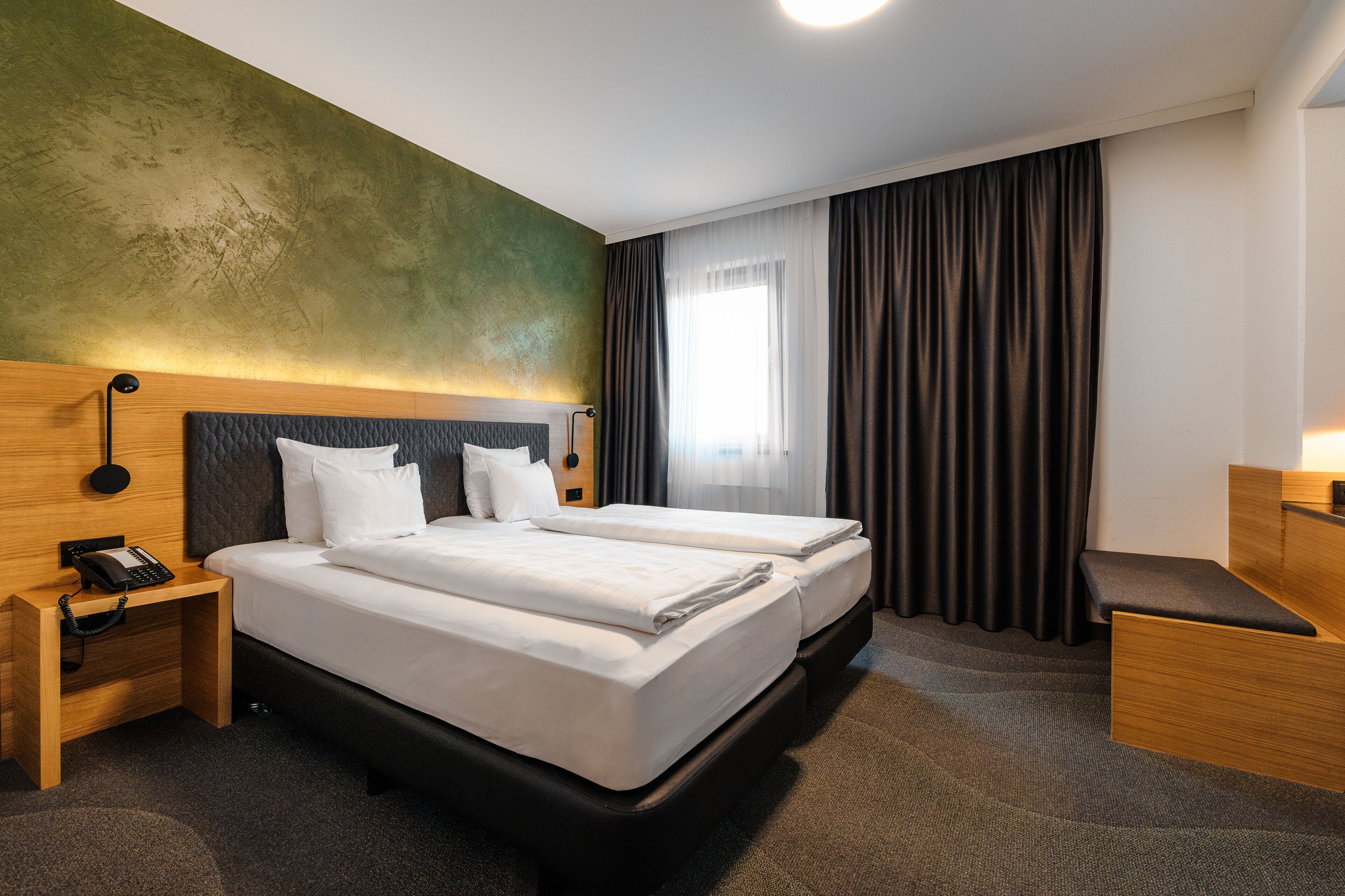 Ein Hotelzimmer mit Doppelbett, Nachttisch mit Telefon, Kopfteil, Fenster mit Vorhängen und Teppichboden.