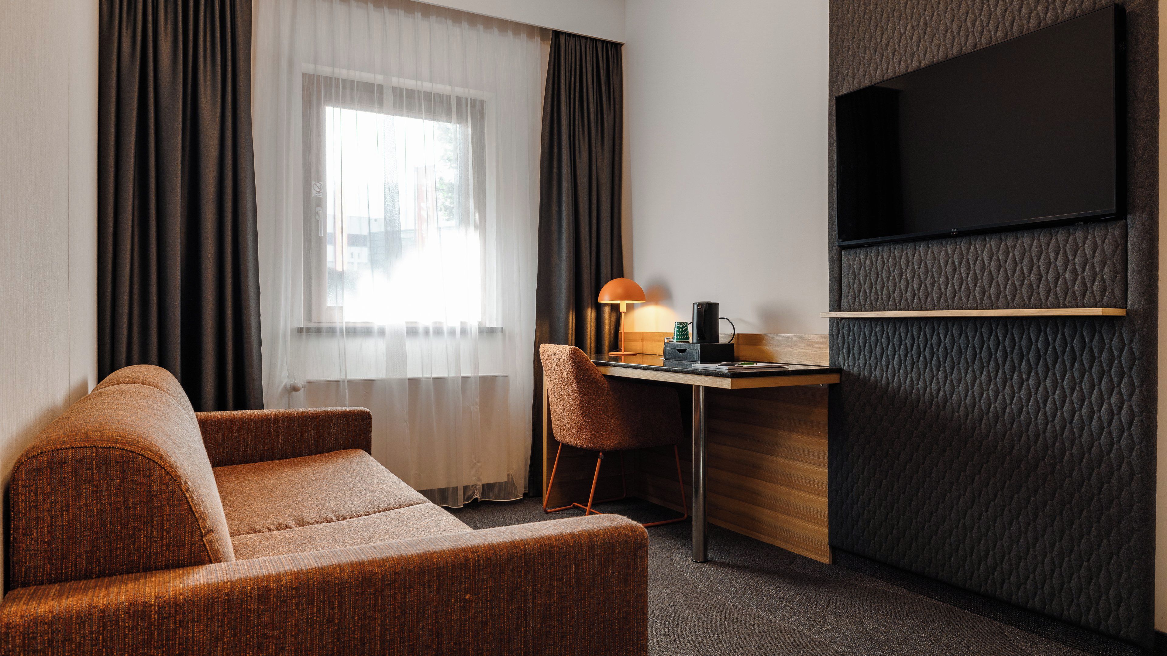 Ein Hotelzimmer mit Sofa, Schreibtisch mit Lampe, Flachbildfernseher und einem Fenster mit Vorhängen.
