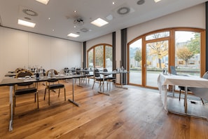 Une salle de conférence équipée de tables, de chaises et d’une grande fenêtre donnant sur des arbres et des immeubles.