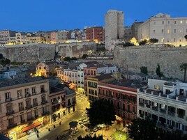 Paesaggio urbano storico con un'imponente muraglia in pietra, edifici tradizionali e una piazza centrale.