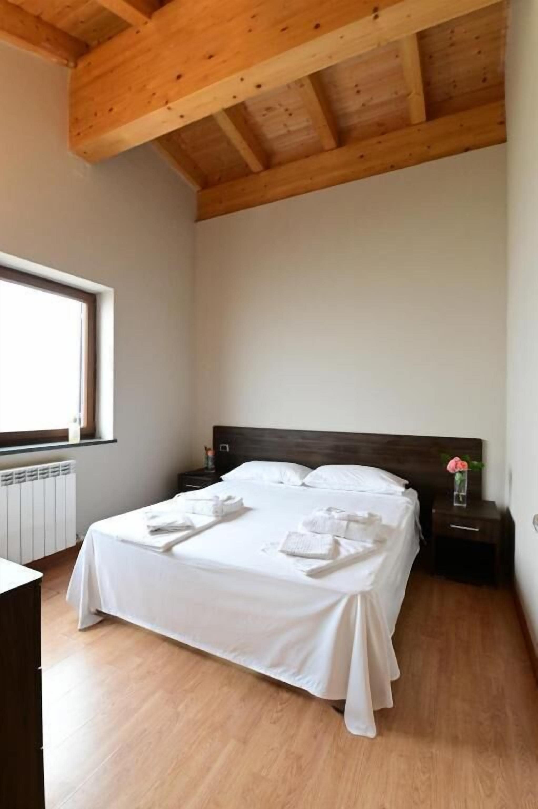 Quarto com teto de madeira, cama com roupa de cama branca, mesa de cabeceira e uma janela.