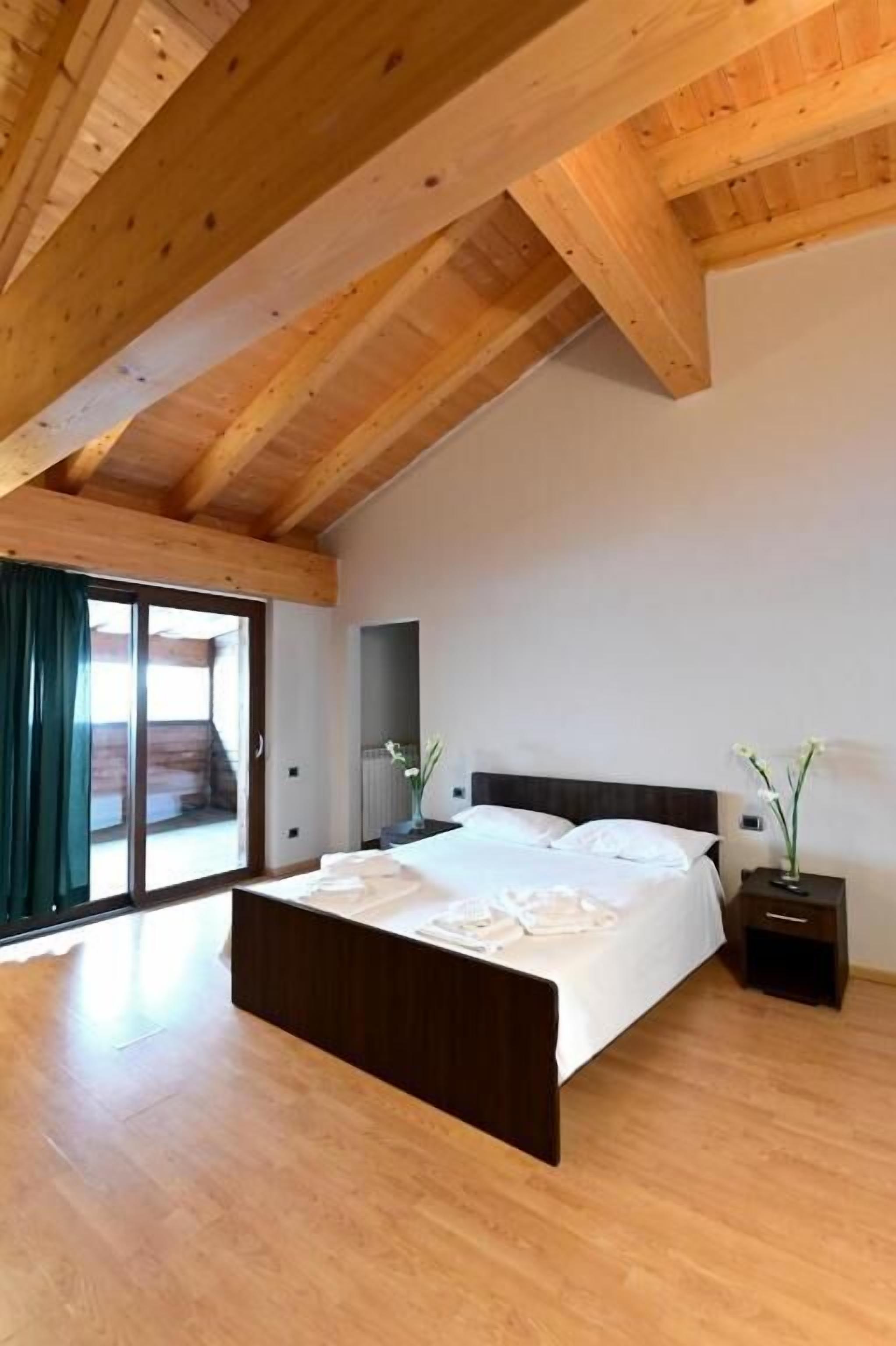 Une chambre avec un plafond en bois, un grand lit, une table de chevet et une vue sur l’extérieur, visible à travers une porte vitrée.