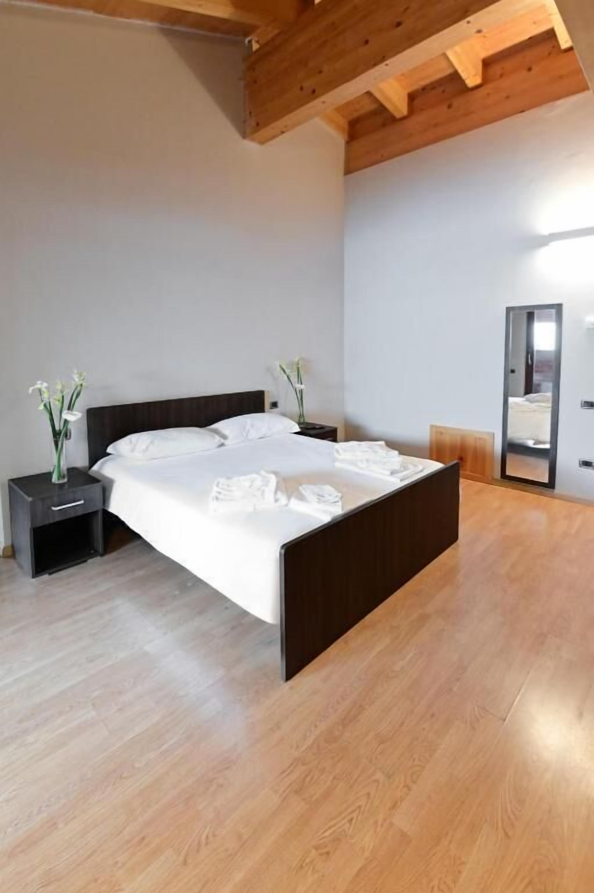 Quarto com piso de madeira, cama com roupa de cama branca, mesa de cabeceira, espelho e vigas de madeira no teto.