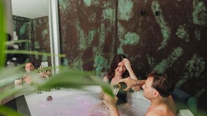 Eine Badewanne, in die eine Person Wasser gießt, ein Spiegel der Szene blickt entgegen und eine strukturierte Wand im Hintergrund.
