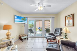 A living room with a ceiling fan, a TV, a sofa, a chair, a lamp, and a view of the beach through the sliding glass doors. - Shalimar 302 (Indian Rocks Beach)