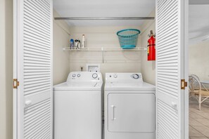 A laundry room with two white front-loading washing machines, a blue laundry basket on a shelf, and a red fire extinguisher on the wall. - Shalimar 302 (Indian Rocks Beach)