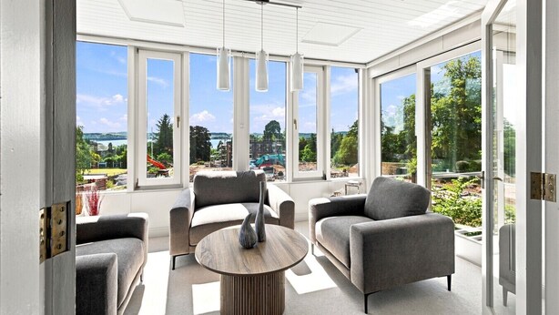Um quarto ensolarado com janelas amplas, uma mesa redonda de madeira e duas poltronas cinzentas.