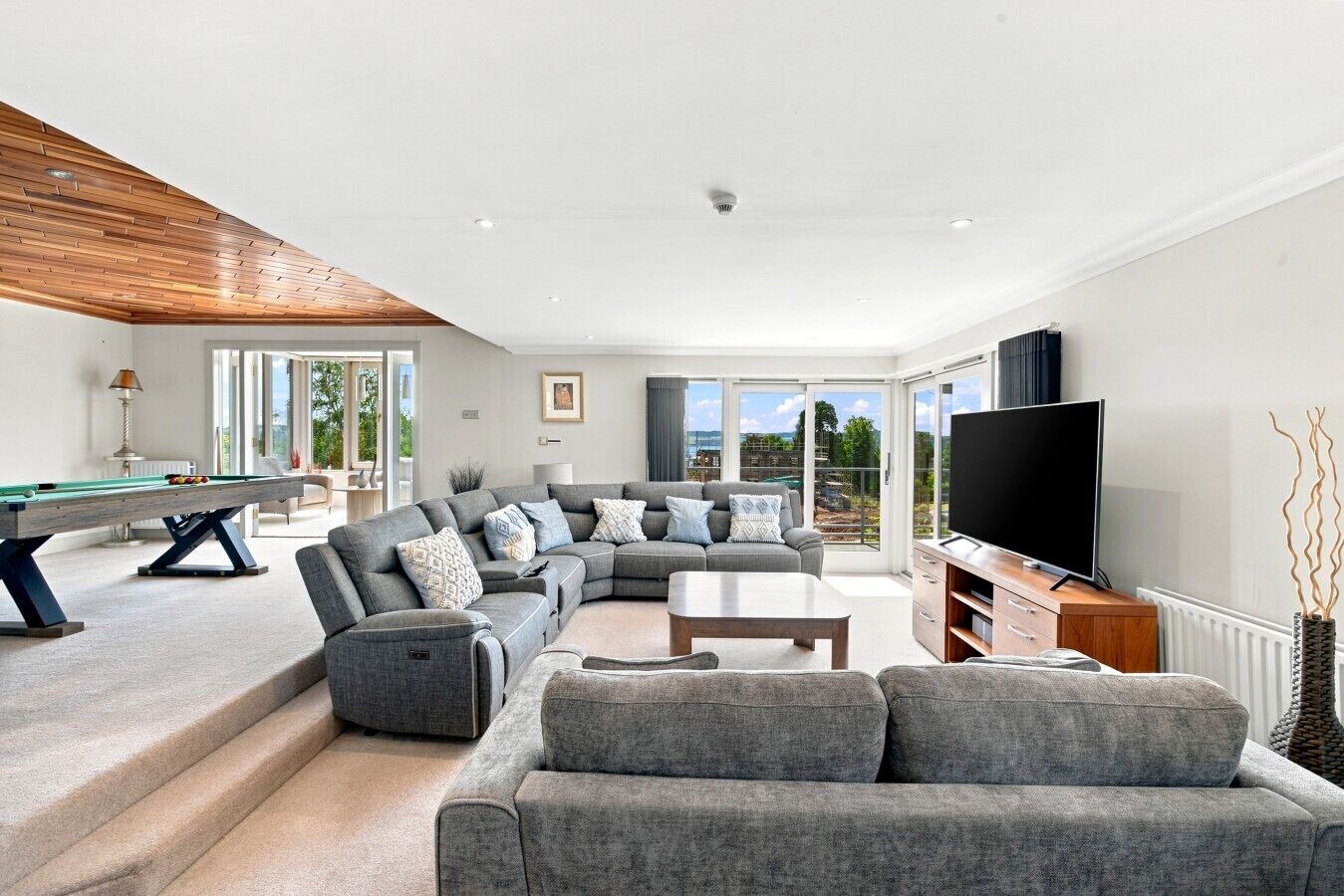 A modern living room with a pool table, grey sofas, a flat-screen TV, and a view of the outdoors.