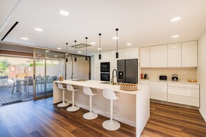A modern kitchen with a central island, bar stools, and a dining area with a glass door leading outside. - Villa Salgados an Golf by Dreamplaces (Albufeira)