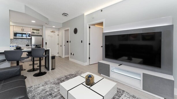 A modern living room with a large flat-screen TV, a dining area with bar stools, and a kitchen in the background. - Staysky Suite Near Universal Pool & Parking (Orlando)