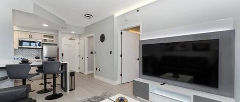 Una sala de estar moderna con un televisor de pantalla plana grande, un comedor con taburetes de bar y una cocina al fondo.
