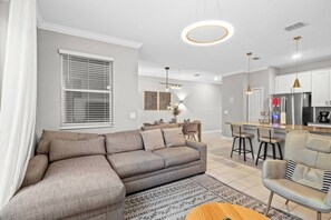 A modern living room with a sectional sofa, a dining area with a small table and chairs, a kitchen with a refrigerator and a coffee machine, and a large window with blinds. - 4BR Storey Lake Home Near Disney With Pool (Kissimmee)