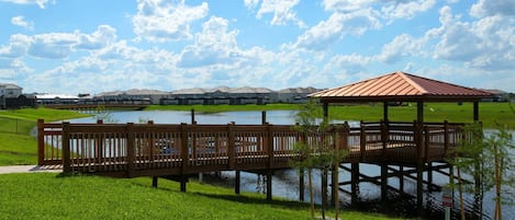 Eine Holzstege über einem See mit einem Pavillon, umgeben von grünem Gras und Wohnhäusern im Hintergrund.