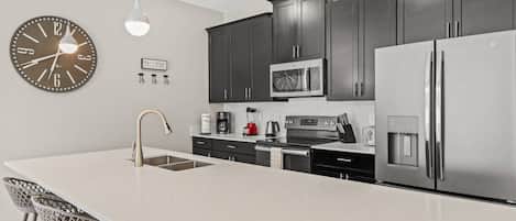 A modern kitchen with dark cabinets, stainless steel appliances, a large island with bar stools, and a wall clock.