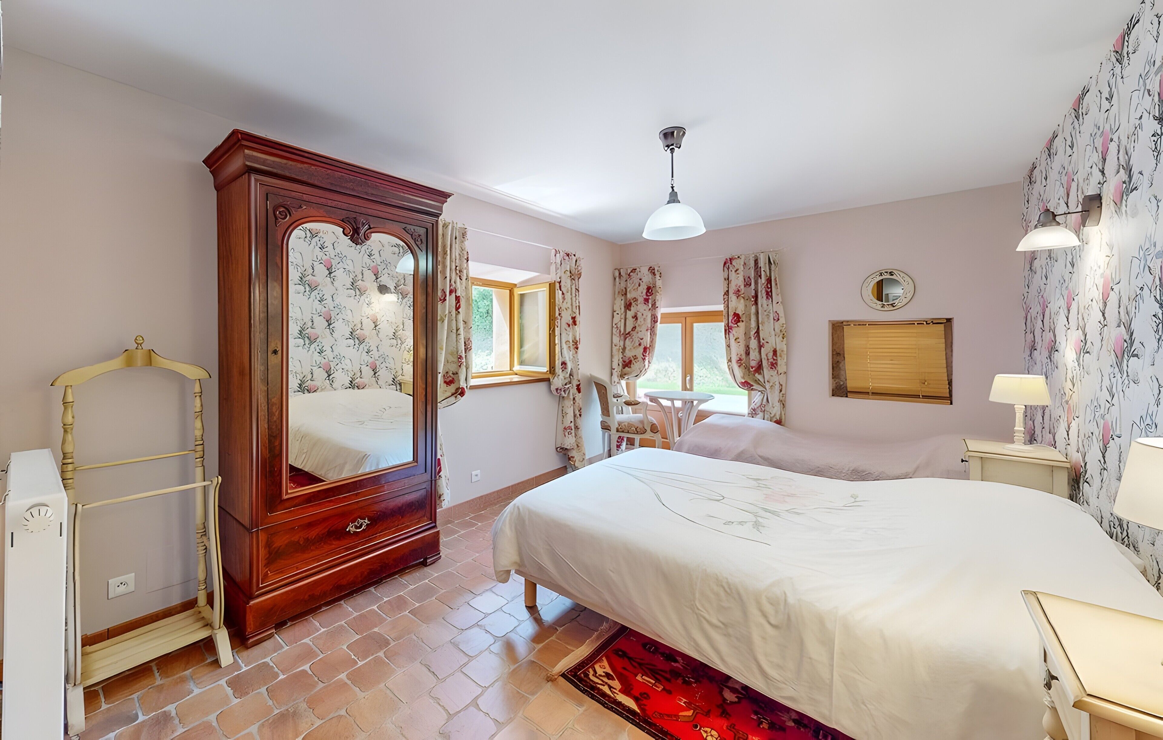 A bedroom with a large bed, a wooden wardrobe, a mirror, a small table, and a chair.
