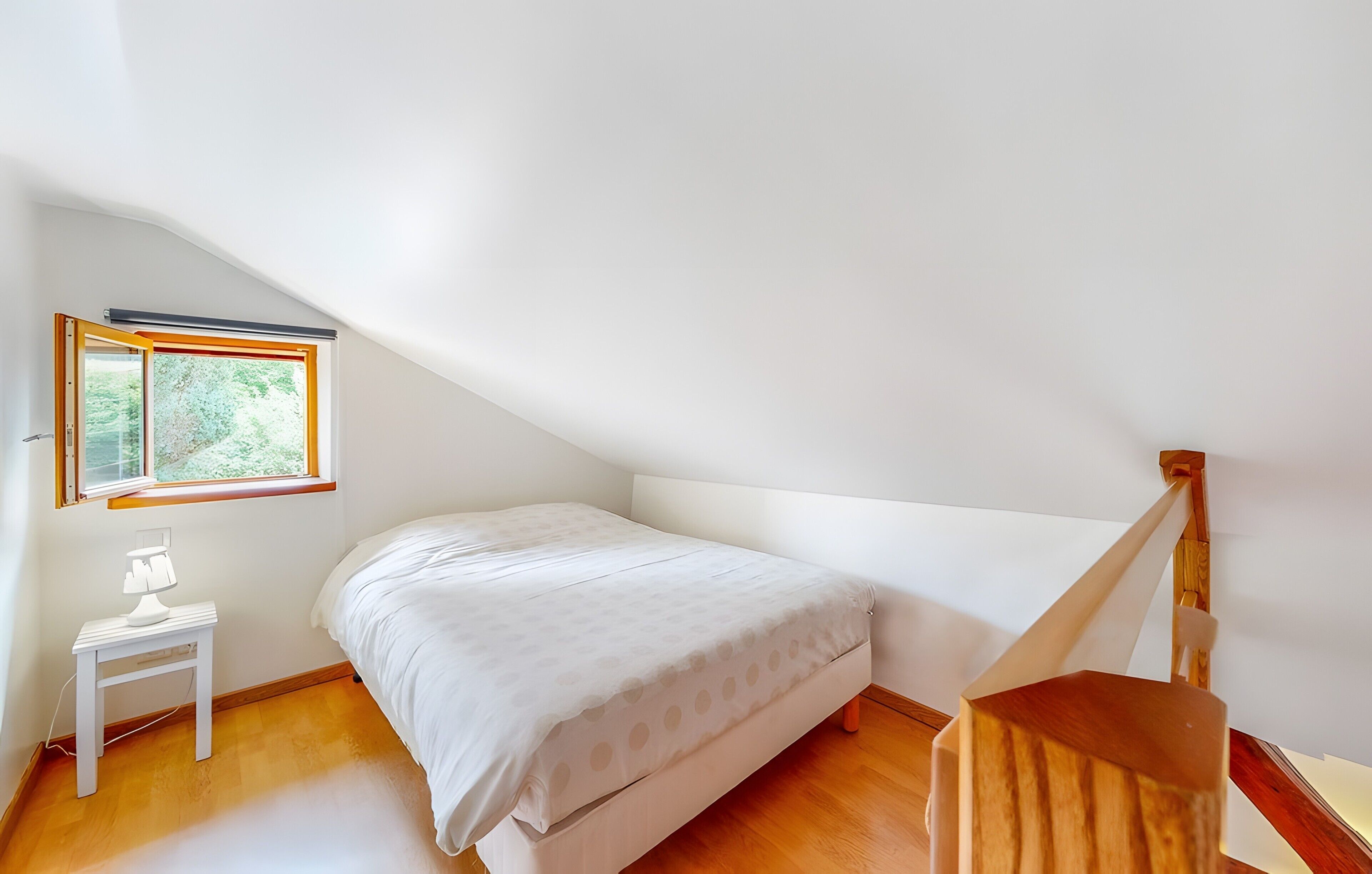 Et soveværelse med en seng, et natbord, en lampe, et vindue og en trappe i træ.