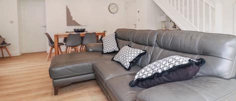 A modern living room with a sectional sofa, a dining table with chairs, a staircase, and a wall clock.
