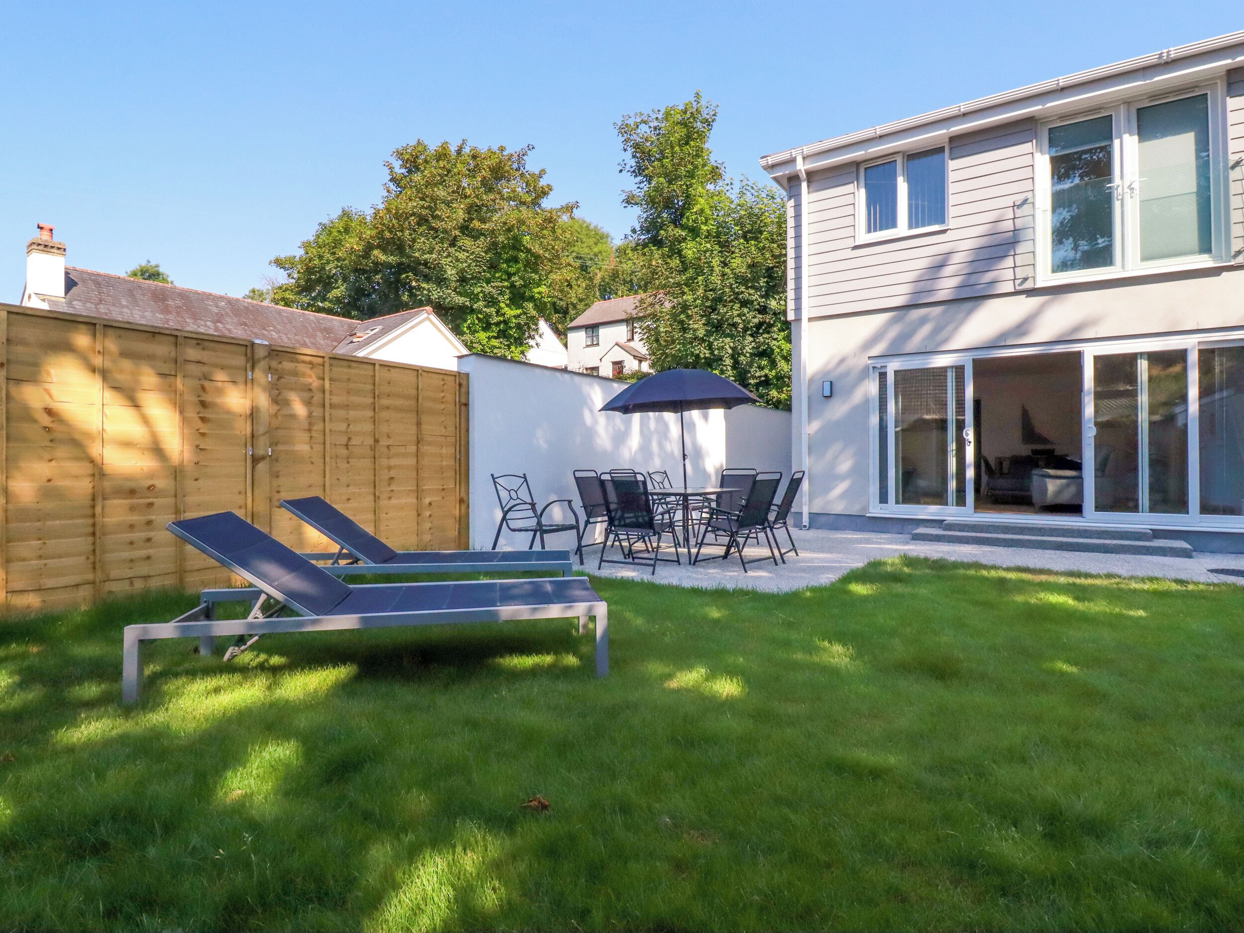 Un jardin avec une pelouse, une clôture en bois, une terrasse dotée d’une table et de chaises, et un bâtiment avec une porte en verre.