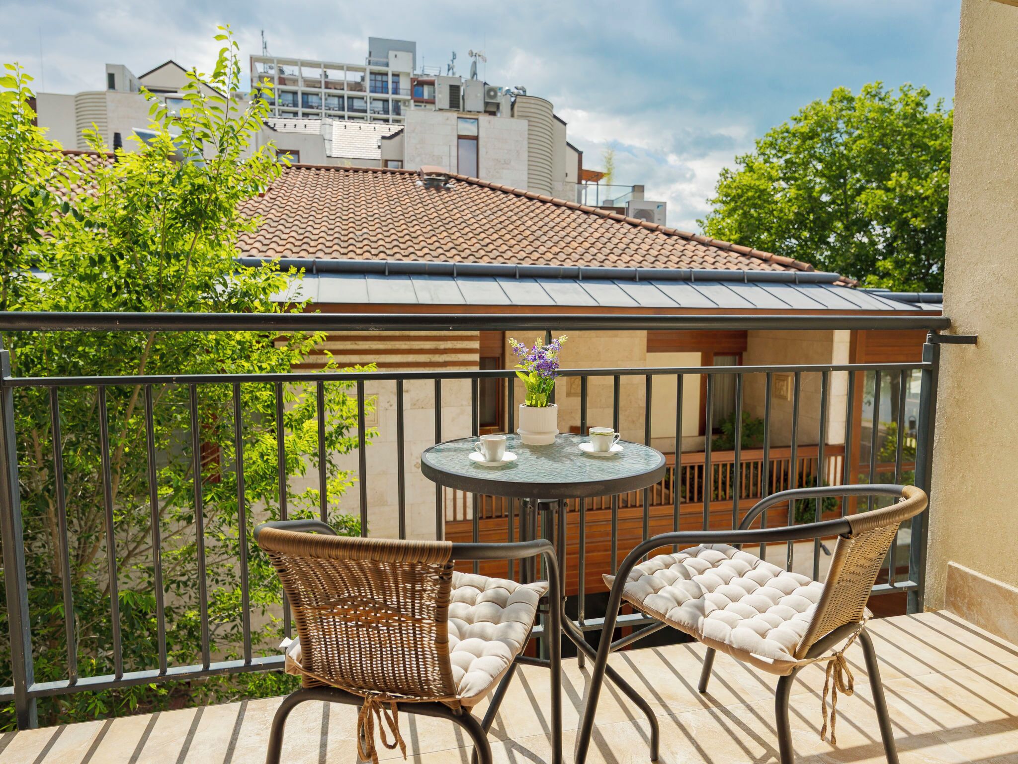 A balcony with two wicker chairs and a small table, a tiled roof, and buildings in the background.