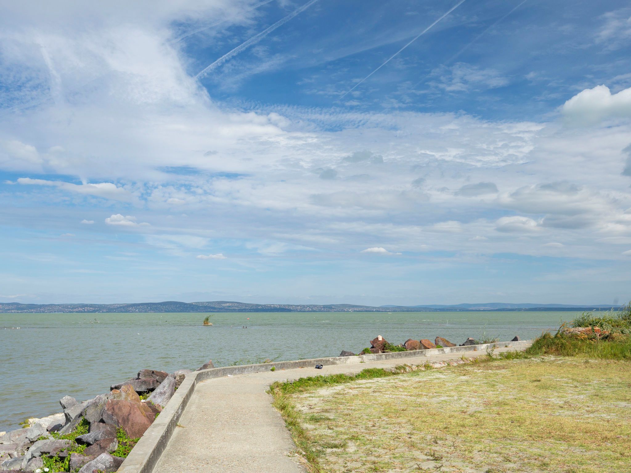 Un paseo marítimo con sendero de hormigón, afloramientos rocosos y vistas al mar y colinas distantes.