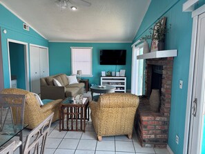 A living room with teal walls, a sofa, wicker chairs, a fireplace, and a television. - The Cozy Coastal, steps away literally from the sand and beach access. (Gulf Shores)
