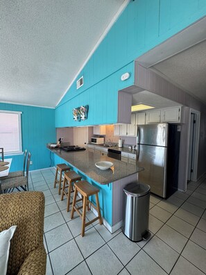 A kitchen with a central island, bar stools, a stainless steel refrigerator, and a microwave. - The Cozy Coastal, steps away literally from the sand and beach access. (Gulf Shores)
