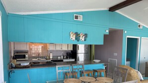 Een keuken met blauwgroene kastjes, een magnetron, een koelkast, en een eethoek met een glazen tafel en rieten stoelen.